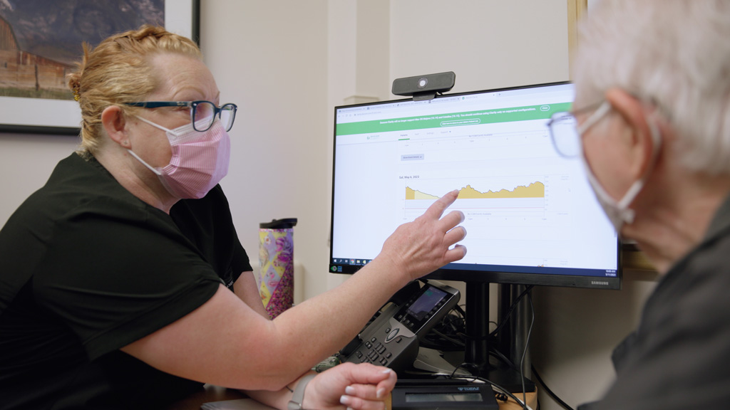 Nurse wearing face mask discusses trends with patient while pointing at a computer monitor.